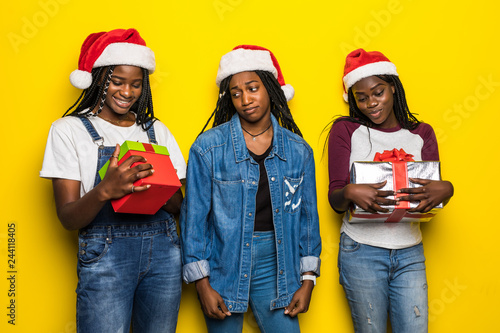 Three young african woman in santa hat share Christmas present on yellow background