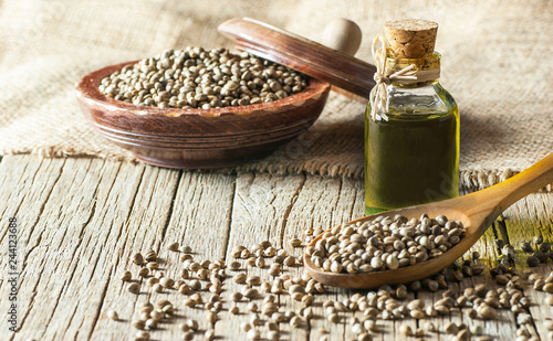 Quadro su tela Heap of dried organic hemp seeds or cannabis plant seeds in spoon and bowl with glass of hemp seed oil on wooden backdrop