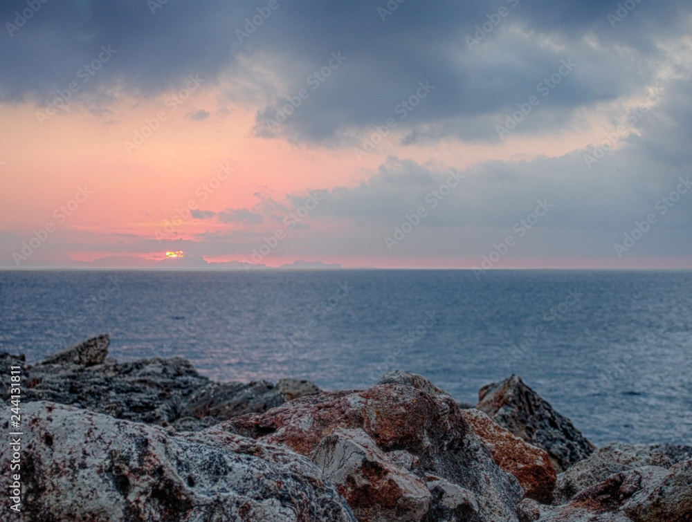 sunset with pink and blue clouds over a calm Mediterranean sea against textured rough coastal rocks in menorca