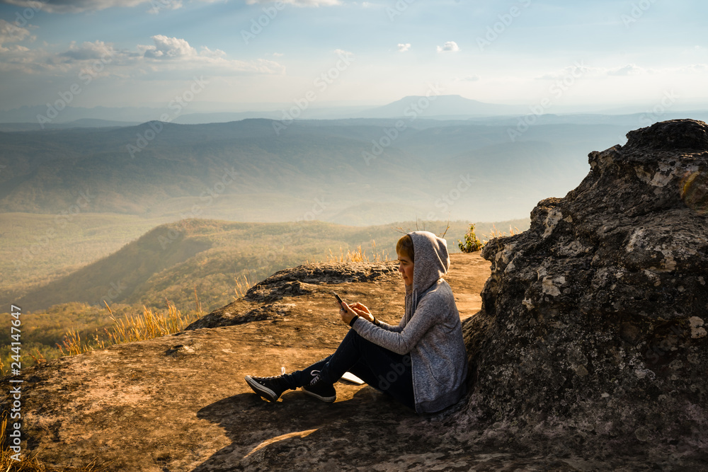A woman sitting on rocky mountain using smartphone and looking out at beautiful natural view