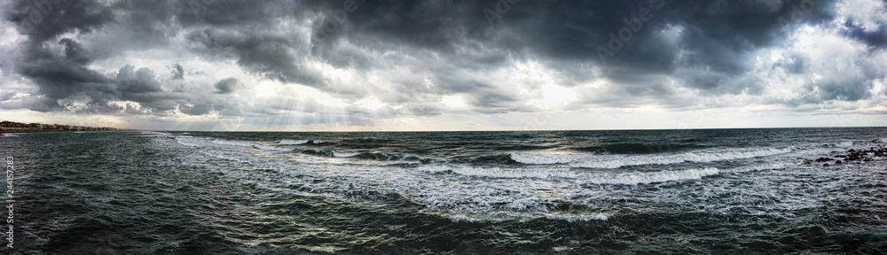 Dramatic weather panoramic view over the sea threatening waves crashing at the shore with overcast sky and sunbeams on the water - find more in my profile