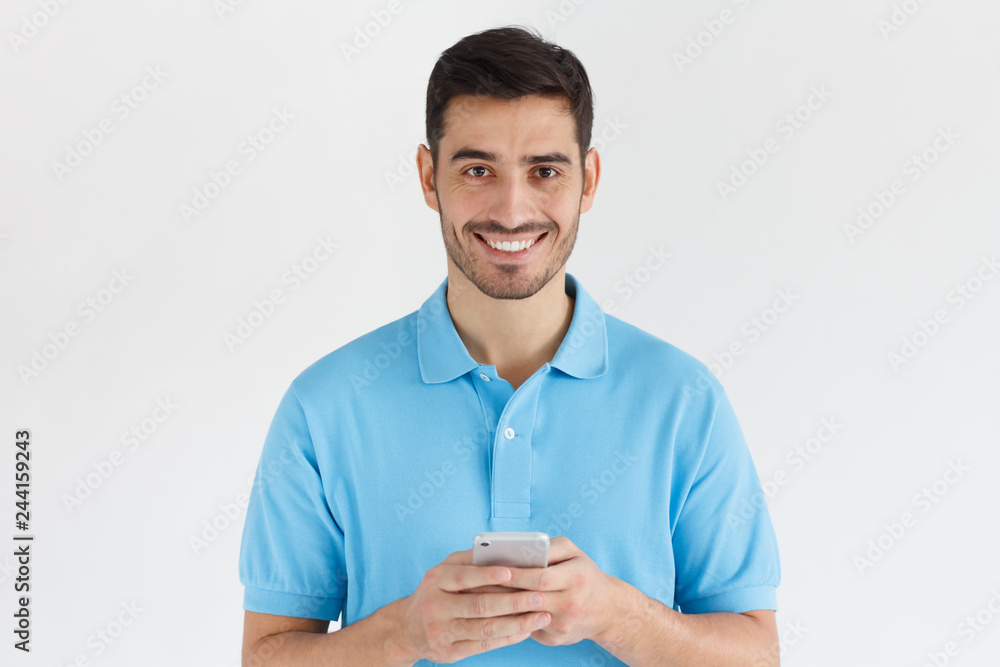 Young man standing isolated on gray background, holding smartphone, looking at camera and smiling nicely Young man standing isolated on gray background, holding smartphone, looking at camera and smiling nicely