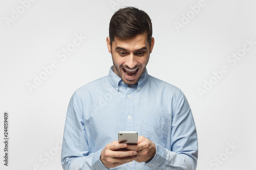 Young businessman looking at smartphone with surprise expression, isolated on gray background