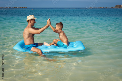 Cute European boy is sitting on a blue floater with his dad. They are happy, smiling and having fun.