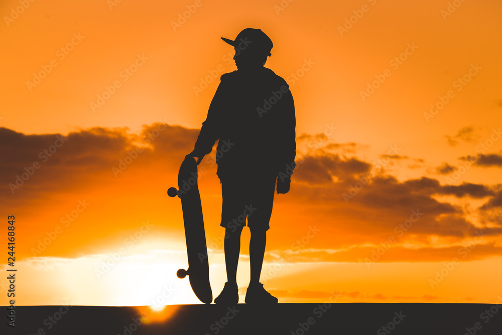Silhouette of young boy plays with skateboard at sunset Backlight of a skater with hat standing in a orange sky with golden reflections on the ocean. Teenager with a skate in hand. Cloudy sky 