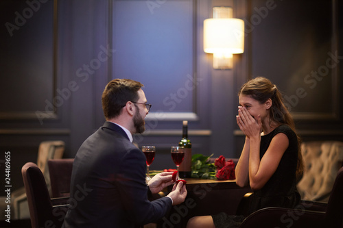 Surprised girl hiding her face in hands while young man making proposal to her in restaurant on valentine day