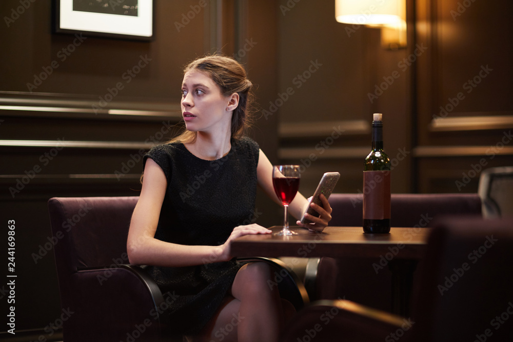 Girl in black dress looking aside while scrolling in smartphone and having  red wine by table in classy restaurant Stock Photo | Adobe Stock