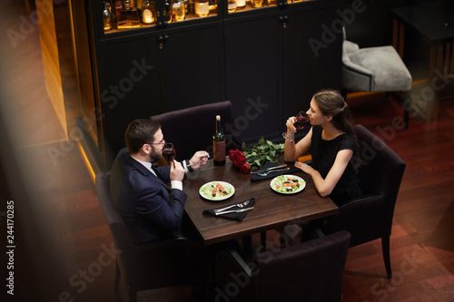 Young amorous dates sitting by table in luxurious restaurant, drinking red wine and talking before eating dinner