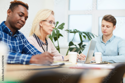 Mature blonde analyst sitting among her young intercultural colleagues or subordinates while networking