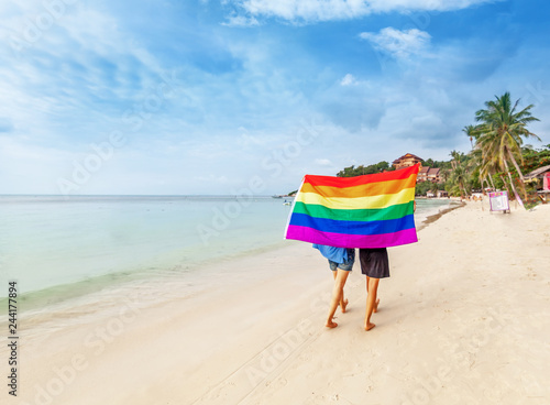 Foto Young lesbian couple walks on a bright tropical beach with a rainbow flag, equal