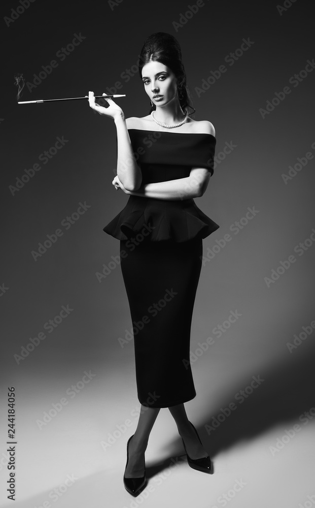 Retro shot: beautiful young woman smoking cigarette. Vintage portrait of  pretty girl in 60s style. Aristocratic elegant lady in black dress and  pearl necklace. Full length, black and white Stock Photo