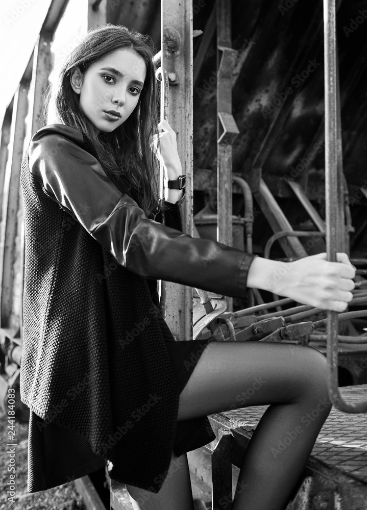 Fashion shot: portrait of beautiful rock girl (informal model) dressed in black jacket and skirt standing near train car. Black and white