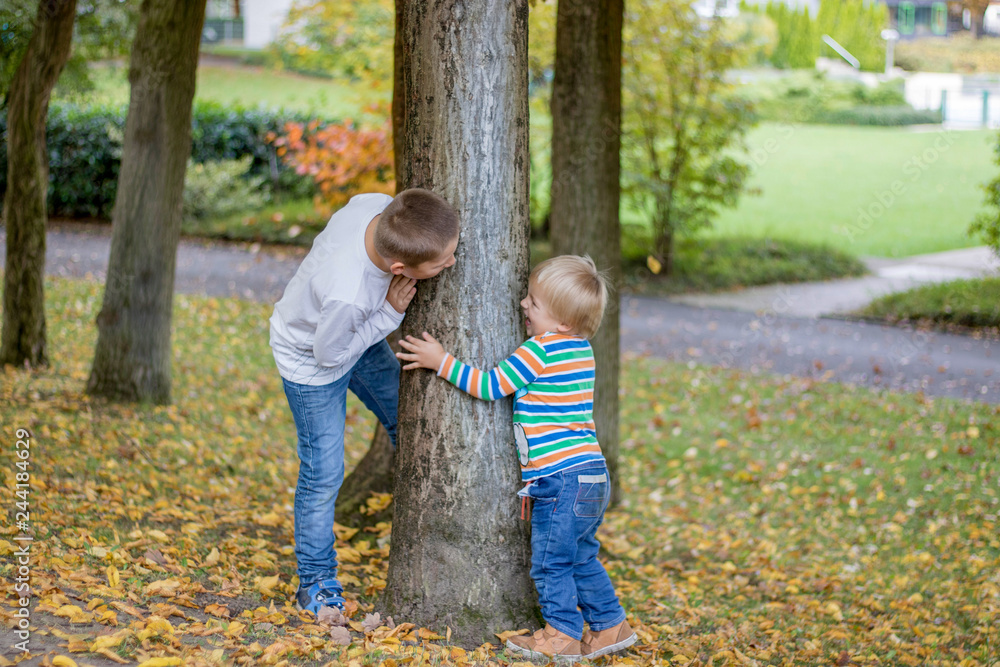 Adorable happy childs, with blond hair peeking around the tree playing hide and seek in a park Adorable happy childs, with blond hair peeking around the tree playing hide and seek in a park