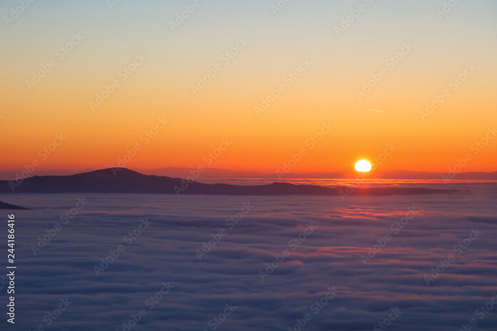 Background photo of low clouds in a mountain valley, red orange sky. Sunrise or sunset view of mountains and peaks peaking through clouds. Winter alpine like landscape of high siberia 