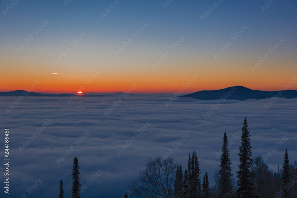 Background photo of low clouds in a mountain valley, red orange blue sky. Sunrise or sunset view of mountains and peaks peaking through clouds. Winter alpine like landscape of high siberia 