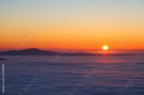Background photo of low clouds in a mountain valley  red orange sky. Sunrise or sunset view of mountains and peaks peaking through clouds. Winter alpine like landscape of high siberia 