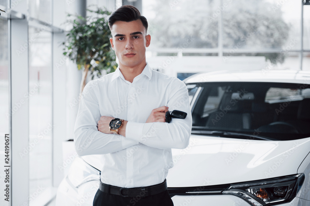 Fototapeta premium A handsome man is a buyer standing next to a new car at the dealer center and looking at the camera.