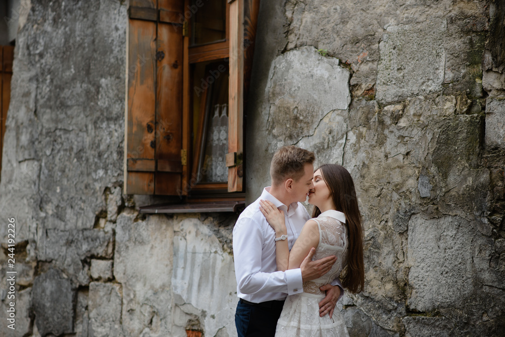 Couple in love is kissing and hugging over the grey urban wall in the morning city. Man and woman are having great time together. Ancient old european city street with stone walls.