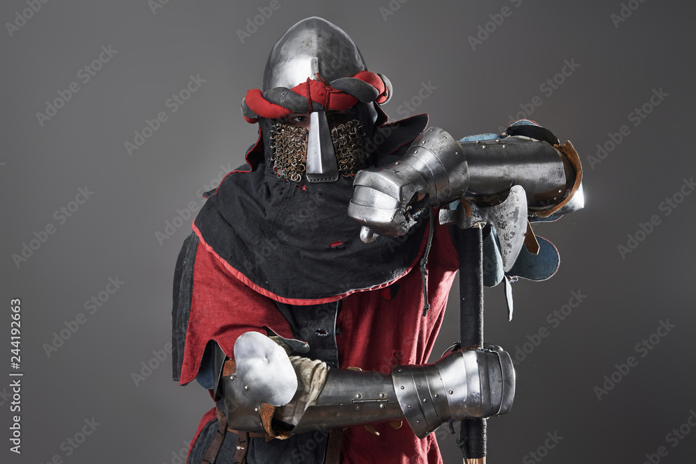 Fototapeta premium Medieval knight on grey background. Portrait of brutal dirty face warrior with chain mail armour red and black clothes and battle axe