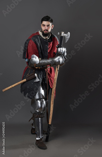 Medieval knight on grey background. Portrait of brutal dirty face warrior with chain mail armour red and black clothes and battle axe