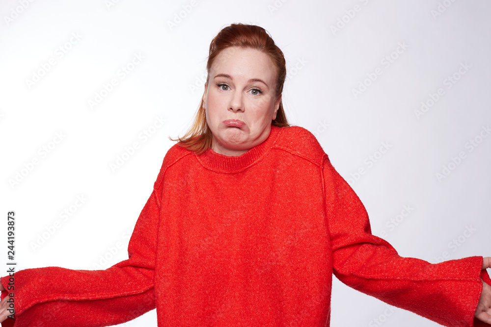 Fototapeta premium People, apathy and attitude concept. Questioned beautiful young woman shrugging with hesitation, feels doubt, has ginger hair, wears casual red oversized sweater, isolated against white background.