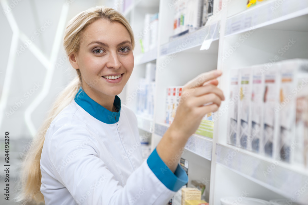 Gorgeous young happy female pharmacist in white labcoat organizing products on shelves, smiling to the camera. Cheerful chemist enjoyign working at her pharmacy. Medicine, health concept Gorgeous young happy female pharmacist in white labcoat organizing products on shelves, smiling to the camera. Cheerful chemist enjoyign working at her pharmacy. Medicine, health concept