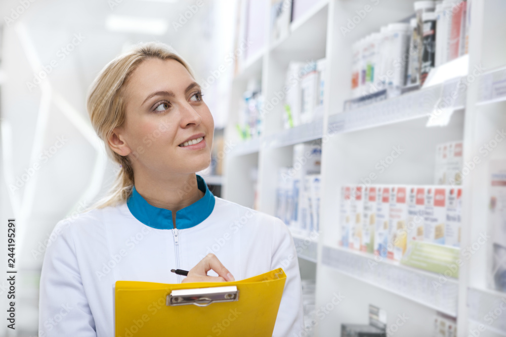 Attractive cheerful female pharmacist checking stock in an aisle, taking notes on her clipboard. Beautiful pharmacist smiling joyfully, working at the local drugstore. Helpful, friendly service concep
