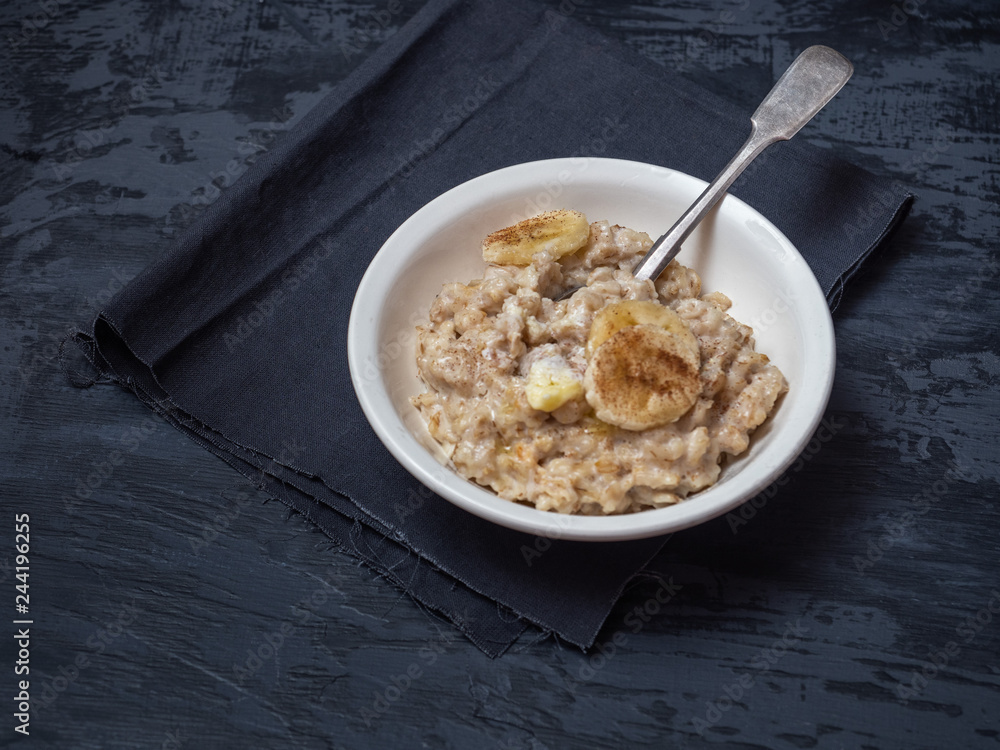 A deep ceramic plate with porridge and a spoon, a sprinkle of cinnamon and banana circles. Under the plate is a dark napkin. A deep ceramic plate with porridge and a spoon, a sprinkle of cinnamon and banana circles. Under the plate is a dark napkin.