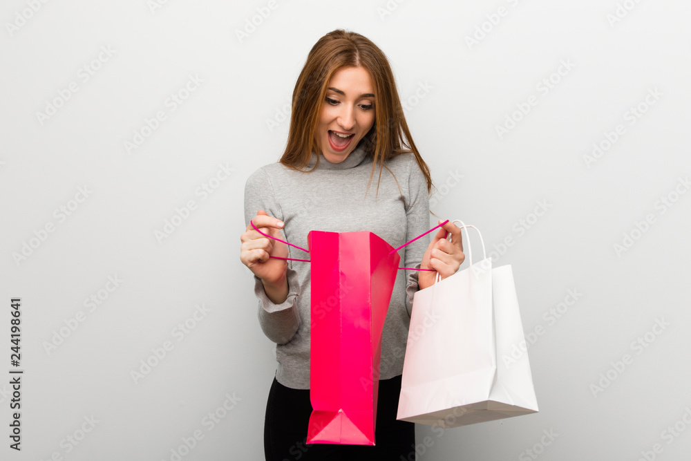 Redhead girl over white wall surprised while holding a lot of shopping bags Redhead girl over white wall surprised while holding a lot of shopping bags