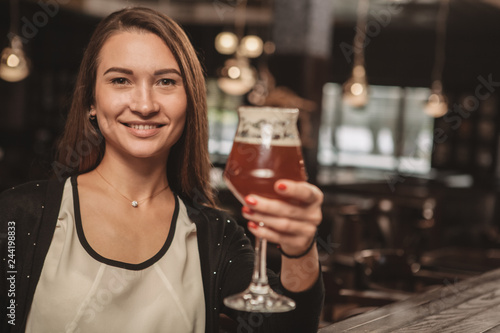 Happy beautiful woman toasting with her glass of delicious craft beer, smiling to the camera, copy space. Attractive female enjoying beer at local brewery restaurant. Celebration, event concept