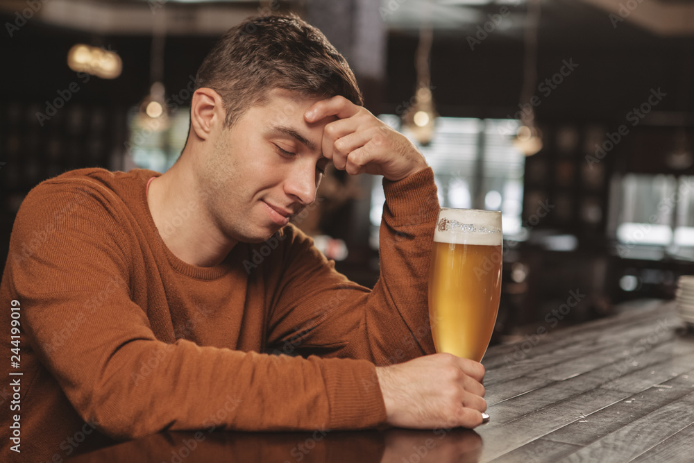 Young drunk man sleeping at the pub counter, smiling joyfully, holding full glass of delicious beer, copy space. Attractive tired man relaxing, drinking beer after long working day. Relaxation concept