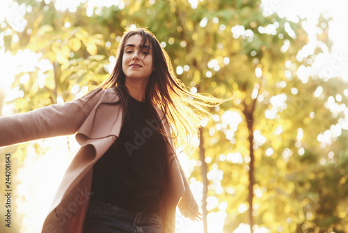 Young happy brunette girl standing and spreading hands wide open, wearing casual coat with autumn yellow trees on the light blurry background. Freedom concept