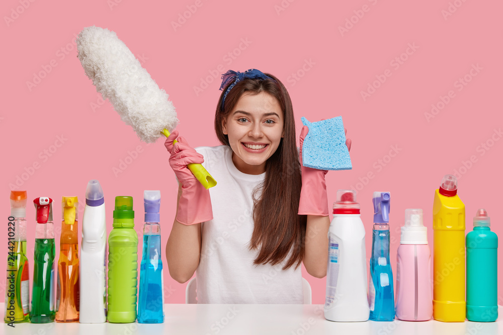 Joyful dark haired woman carries rag and brush, smiles happily, dressed in casual clothes, sits at white desk with cleaning products, isolated over pink background. Happiness, housekeeping concept - obrazy, fototapety, plakaty 