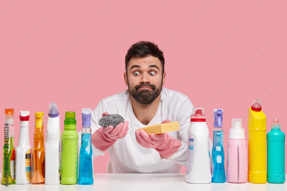 Φωτογραφία Indoor shot of surprised bearded man looks with eyes popped out, wears casual clothes, uses different detergents, isolated over pink background, prepares for washing dish