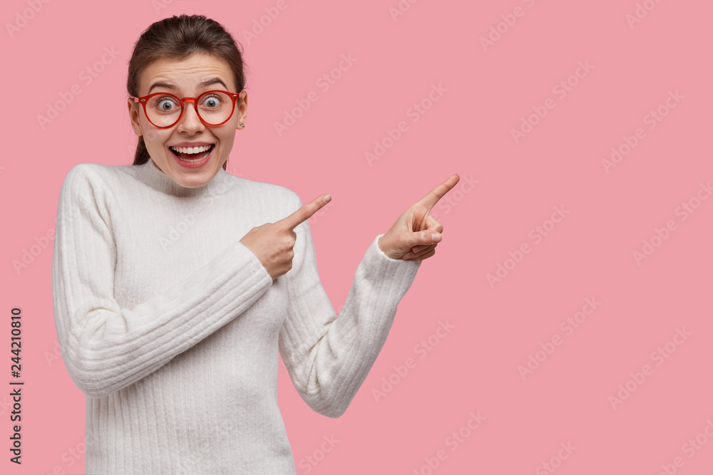 Studio shot of joyful woman points right with finger gun gesture, shows something wonderful against pink background, dressed in white casual outfit, isolated over pink studio wall. Check this - obrazy, fototapety, plakaty 