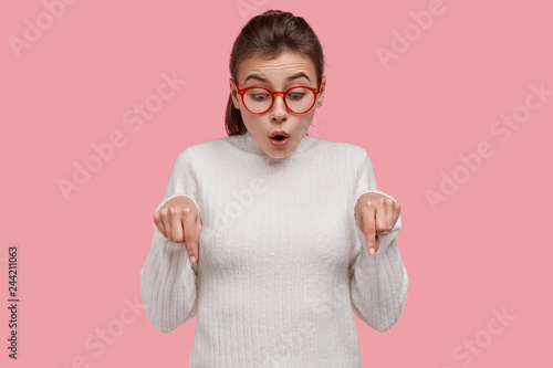 Horizontal view of amazed dark haired young woman with pony tail, points down with fore fingers, openes mouth from amazement, dressed in casual clothes, models over pink background. Studio shot photo