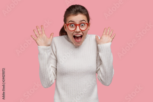 Photo of overemotive young woman reacts on sudden fake news, shows palms, keeps jaw dropped, wears transparent spectacles, dressed in white jumper, models over pink background. Emotions concept photo