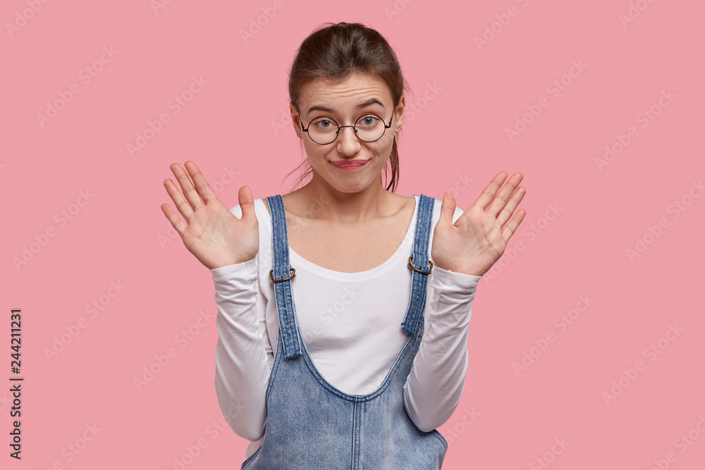 Waist up shot of dissatisfied Caucasian girl shows palms towards camera, demonstrates stop gesture, looks with apathy, wears round spectacles and denim clothes, isolated over pink background Waist up shot of dissatisfied Caucasian girl shows palms towards camera, demonstrates stop gesture, looks with apathy, wears round spectacles and denim clothes, isolated over pink background