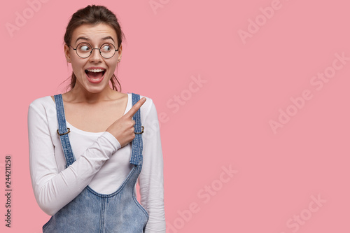 Photo of pleased brunette woman with joyful expression, indicates at upper right corner, wears optical glasses and denim outfit, shows free space for your promotional content. Wow, wonderful sales