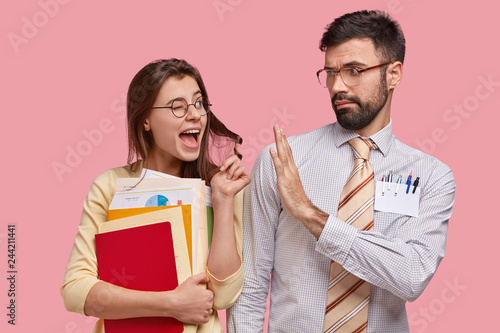 Studio shot of positive woman blinks eyes, flirts with colleague or classmate who refuses have relationships, shows no gesture, dressed in formal clothes, stand closely to each other, isolated on pink