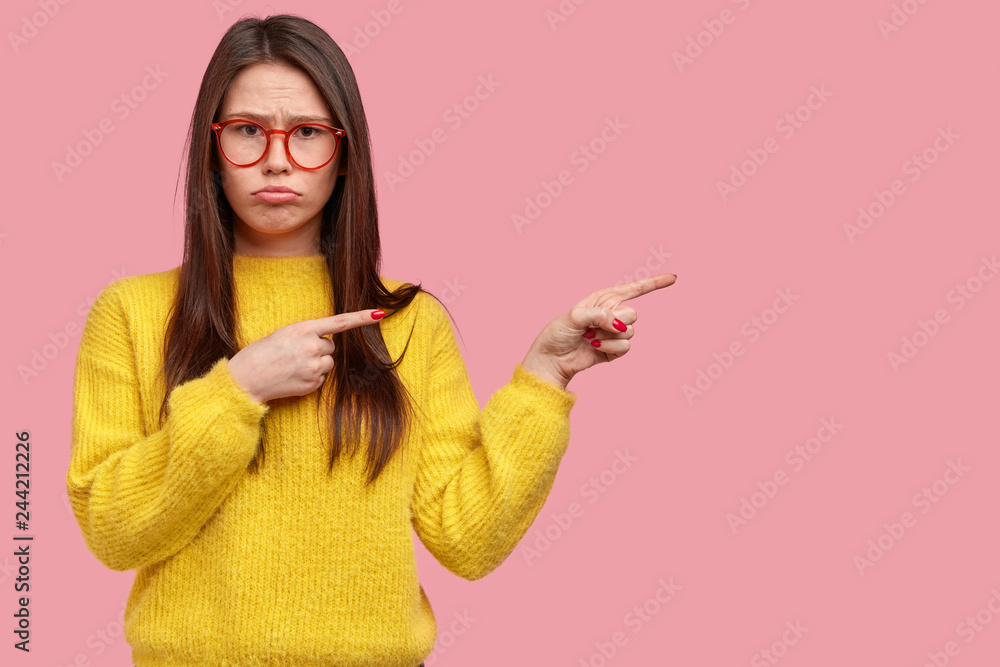 Sad European woman has dejected facial expression, doesnt like something, points aside with both index fingers, dressed in casual yellow jumper, isolated over pink background with blank space Sad European woman has dejected facial expression, doesnt like something, points aside with both index fingers, dressed in casual yellow jumper, isolated over pink background with blank space