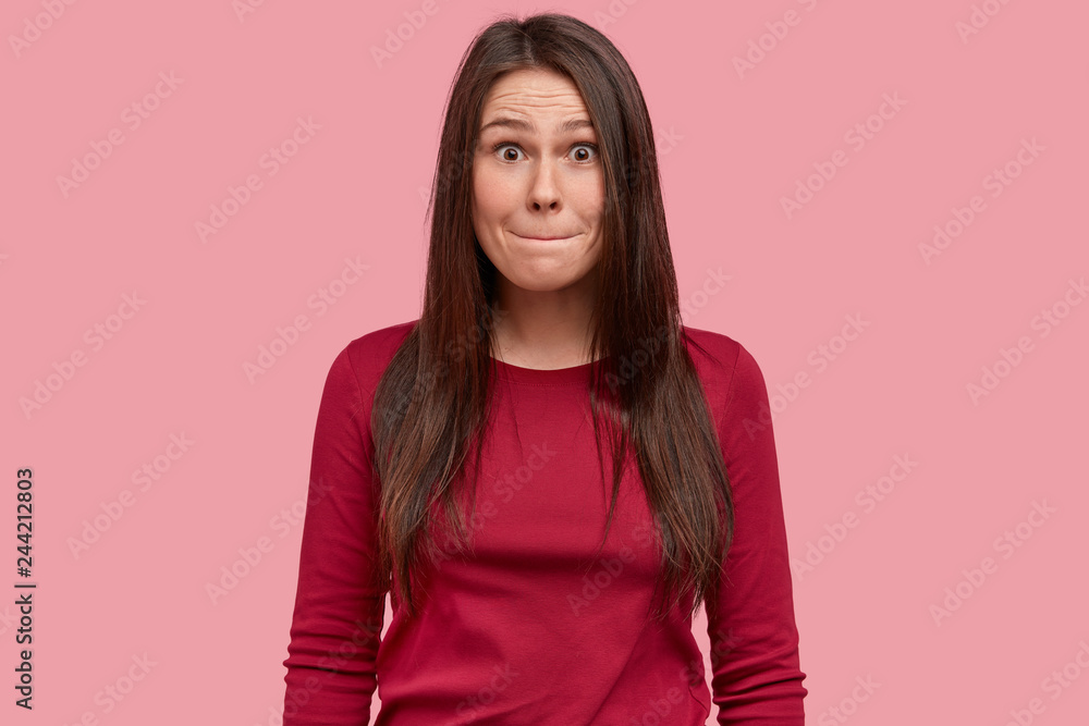 Isolated shot of attractive young woman with long black hair, presses lips, has surprised expression, wears causal red clothes, poses over pink background, reacts on something, feels puzzled Isolated shot of attractive young woman with long black hair, presses lips, has surprised expression, wears causal red clothes, poses over pink background, reacts on something, feels puzzled