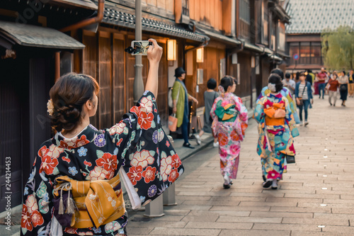 Fotografie, Tablou Japanese girl in kimono taking a photo of a traditional street with wooden house