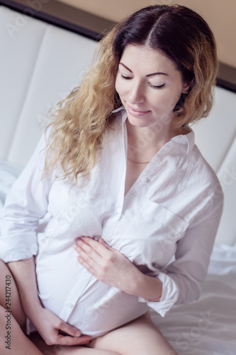 Attractive pregnant woman is sitting in bed and holding her belly