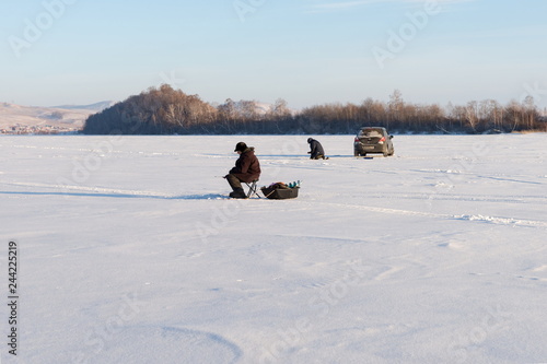 Two fishermen catch fish from the holes on a frozen lake on the background of the island.