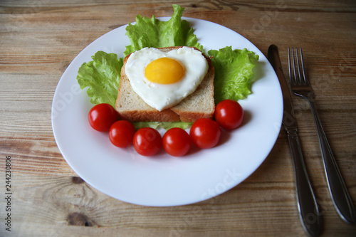 fried eggs with a yellow yolk in the form of a heart on bread, decorated with green salad and red cherry tomatoes on a white plate and wooden rustic background with space for text 
