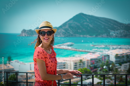 Happy tourist woman with straw sunhat looking to the mediterranean sea and enjoying the blue and scenic seascape in Altea, Alicante, Spain. Living coral dress, color of the year