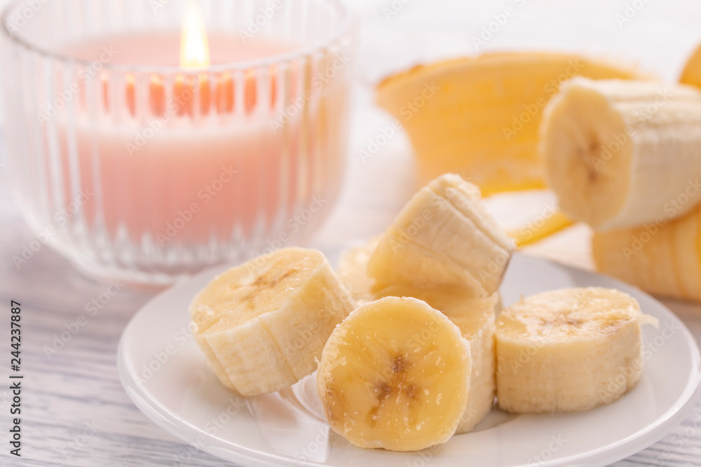 Sliced banana on a white plate and a light wooden table. Pink burning candle nearby. Sliced banana on a white plate and a light wooden table. Pink burning candle nearby.