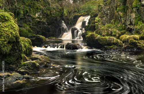 Long exposure shot of Linn Jaw Waterfall, near Livingston, Scotland, with white foam spiral swirls in the water off the waterfall and framed by green mossy rocks. West Lothian. UK. Nature. Landscape photo