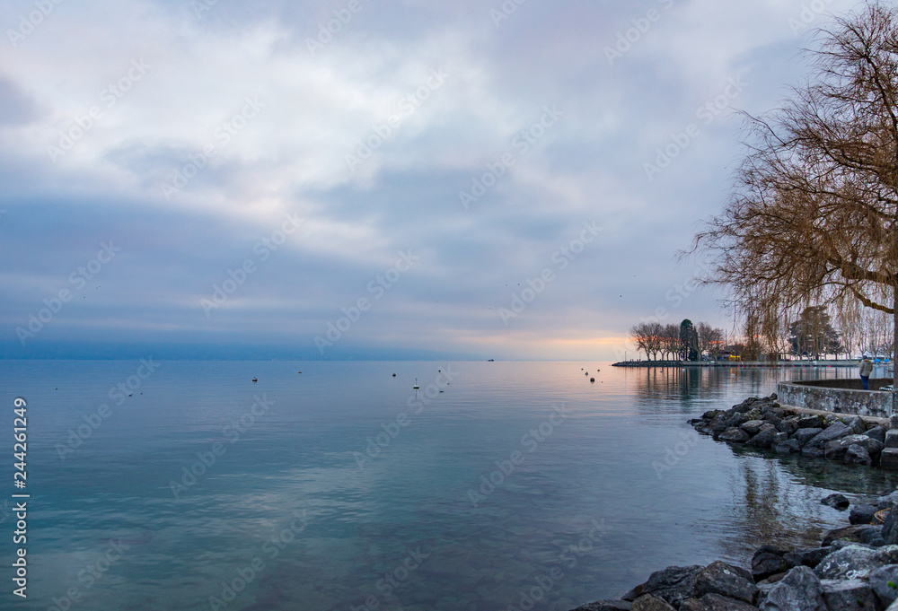 Outdoor scenery of beautiful tranquil natural walkway and promenade along lakeside of lake Geneva and background of misty, cloudy and twilight sky over water in Lausanne, Switzerland in winter.